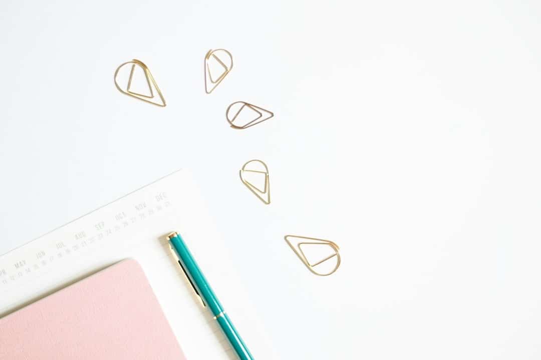 Gold paper clips scattered on a white desk with a pink planner and teal pen.